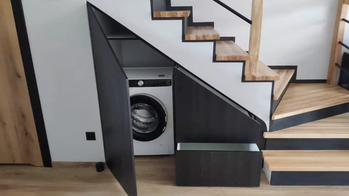 Biała pralka ukryta pod schodami w zabudowie z ciemnego drewna, otwarte drzwiczki szafki i wysunięta szuflada.