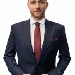 Portret mężczyzny w granatowym garniturze, białej koszuli i bordowym krawacie, z poszetką w kieszonce, na białym tle.