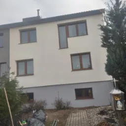 Ocieplony, dwukondygnacyjny dom z jasną elewacją i brązowymi ramami okiennymi, widoczny fragment posesji z krzyżem i ścieżką z kostki brukowej, obok sterta śmieci w czarnych workach.