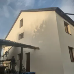 Elewacja domu po tynkowaniu, widoczny cień drzewa na świeżo nałożonym tynku, pracownik na schodach zabezpiecza wejście do budynku.
