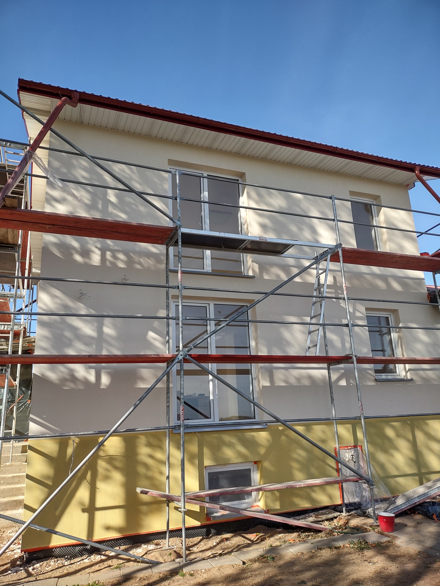 Dwukondygnacyjny dom z elewacją w trakcie prac wykończeniowych, widoczne rusztowania, dolna część ściany w kolorze żółtym, górna w odcieniach beżu, okna w białych ramach.
