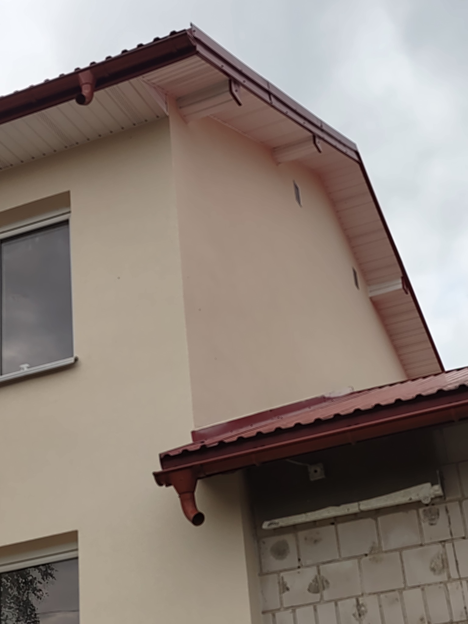 Fragment elewacji domu z beżowym tynkiem, czerwonymi rynnami i częściowo wykończoną ścianą z bloczków betonowych.