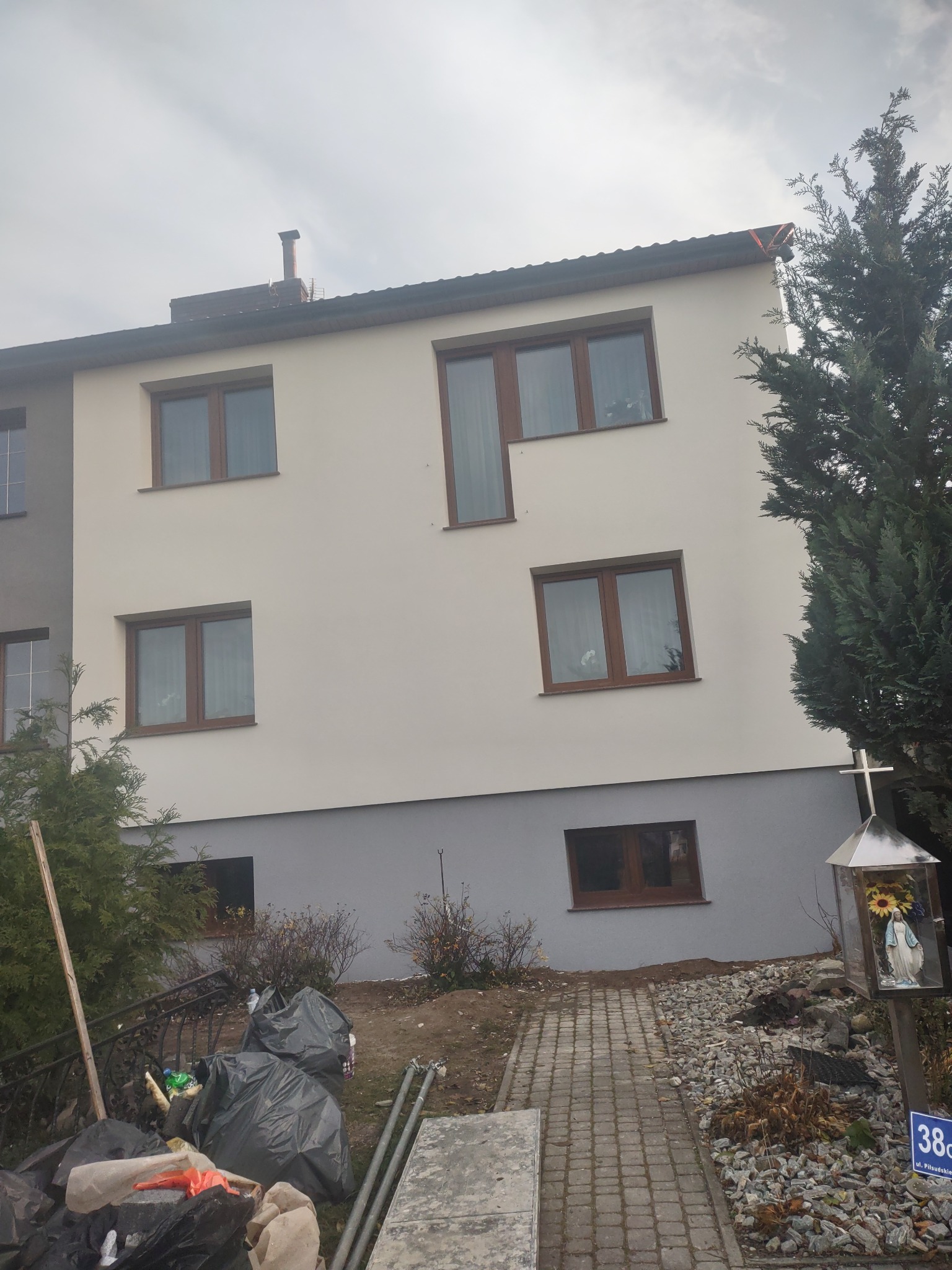 Ocieplony, dwukondygnacyjny dom z jasną elewacją i brązowymi ramami okiennymi, widoczny fragment posesji z krzyżem i ścieżką z kostki brukowej, obok sterta śmieci w czarnych workach.