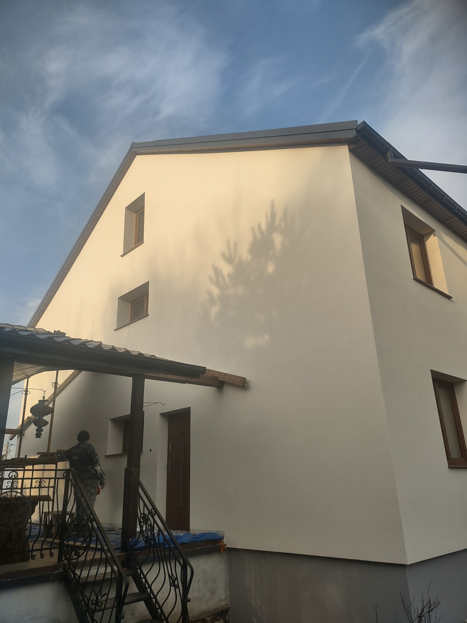 Elewacja domu po tynkowaniu, widoczny cień drzewa na świeżo nałożonym tynku, pracownik na schodach zabezpiecza wejście do budynku.