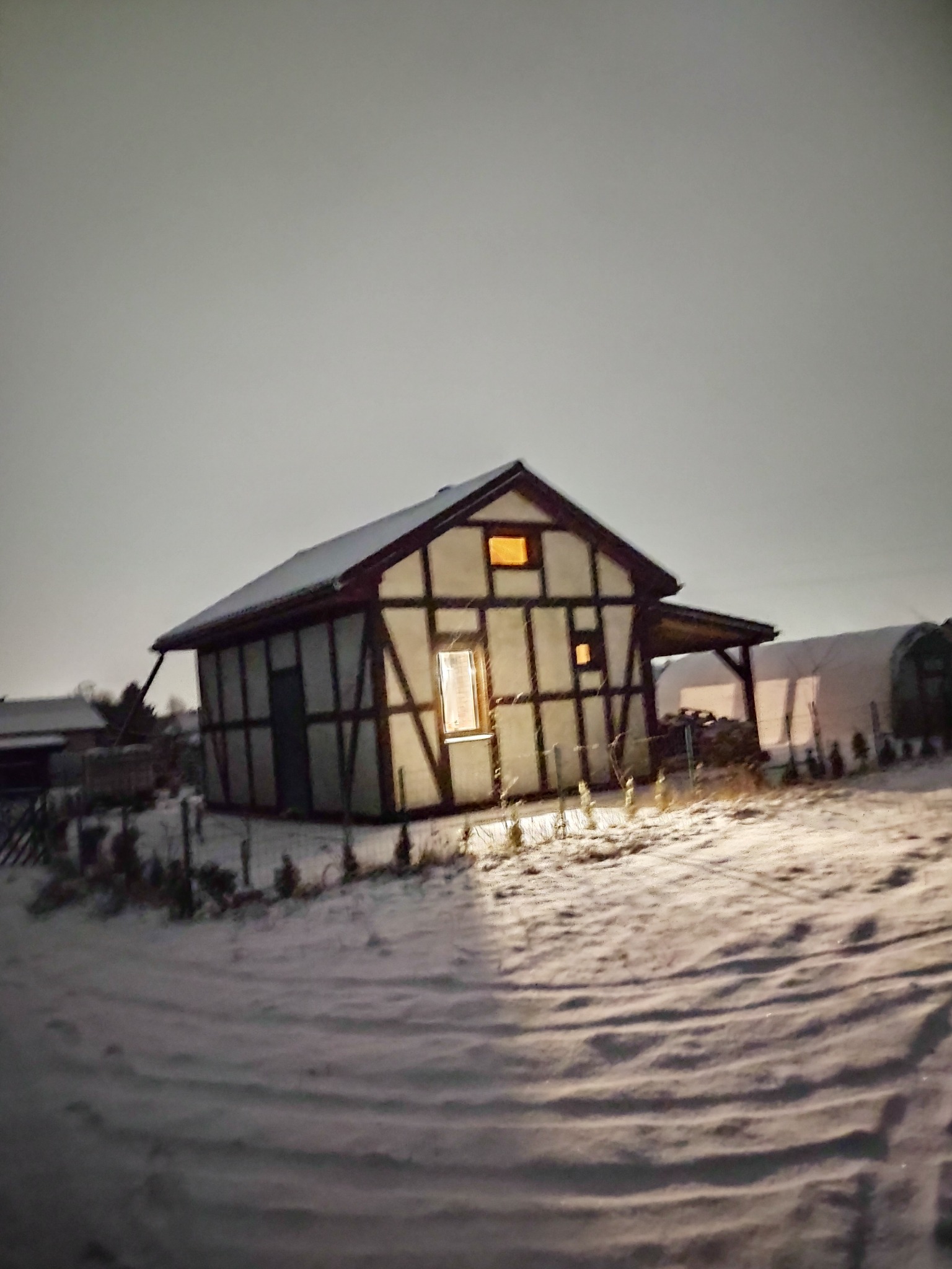 Zimowy krajobraz z widokiem na dom w konstrukcji szkieletowej, oświetlony od wewnątrz, pokryty śniegiem, z widocznymi śladami na śniegu i szklarnią w tle.