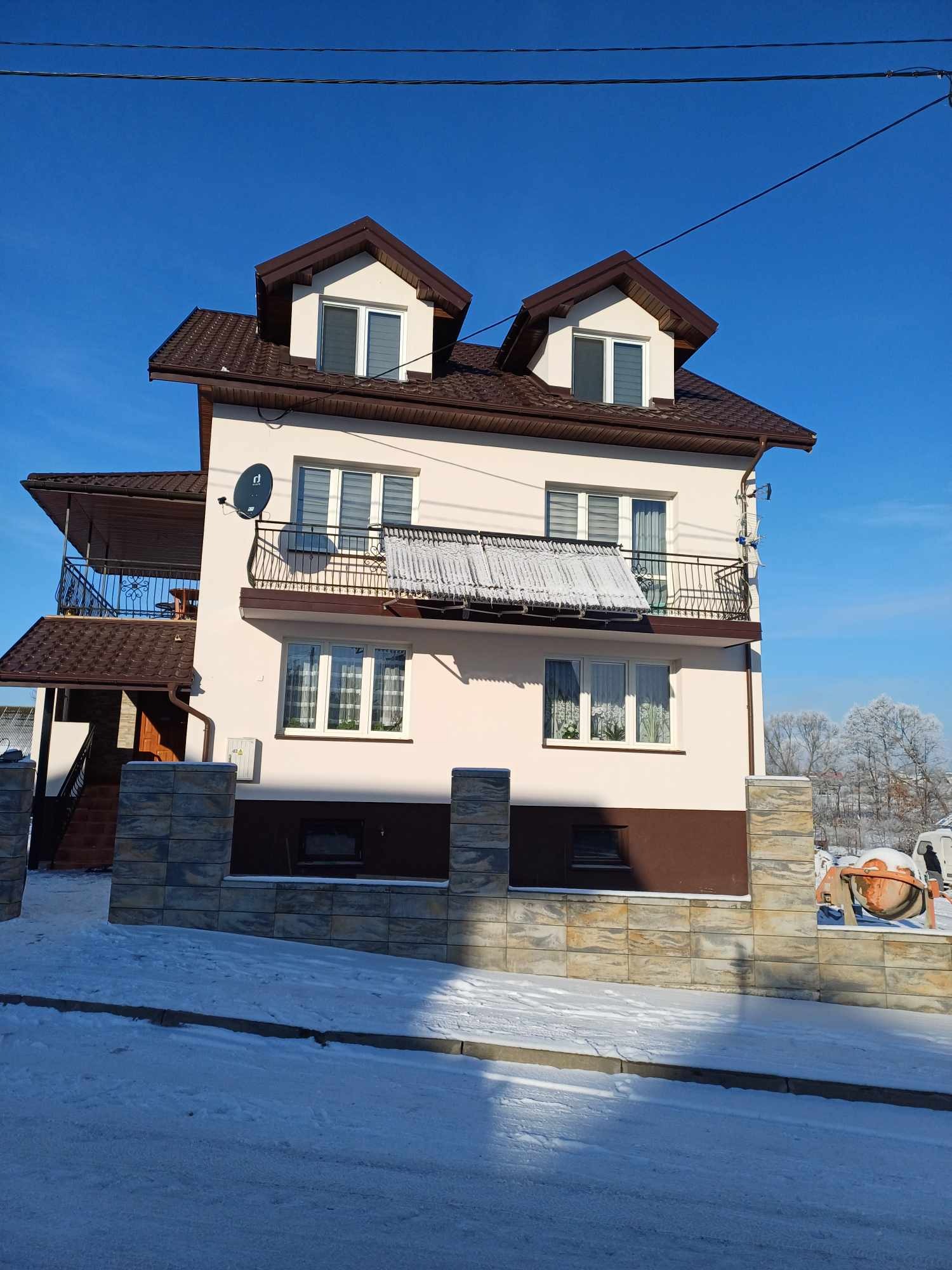 Dwupiętrowy dom z brązowym dachem i elewacją w odcieniach beżu, widok z zewnątrz w zimowy dzień, śnieg na chodniku i ogrodzeniu z kamiennych bloków, antena satelitarna na elewacji.