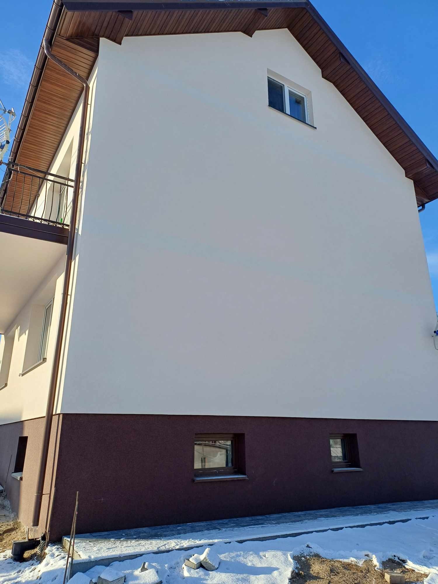 Dwukondygnacyjny dom z nową, jasną elewacją i ciemnobrązowym cokołem, widoczny balkon z kutą balustradą, fragmenty śniegu przy fundamentach.