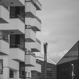 FORMU Mikołaj Małecki - Czarno-białe zdjęcie fragmentu nowoczesnego budynku mieszkalnego z balkonami, z widocznym budynkiem biurowym w tle i elementami oświetlenia ulicznego.