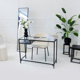 FORMU Mikołaj Małecki - Minimalistyczne wnętrze: szklane biurko z czarną ramą, kremowe krzesło, lustro w czarnej ramie, kwiaty w wazonie i roślina doniczkowa na tle białej ściany.