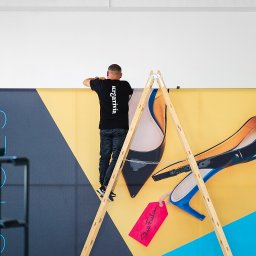 FORMU Mikołaj Małecki - Mężczyzna na drabinie montuje duży baner reklamowy z grafiką butów na tle żółto-niebieskim. Baner w trakcie instalacji, widoczne narzędzia i drabina.