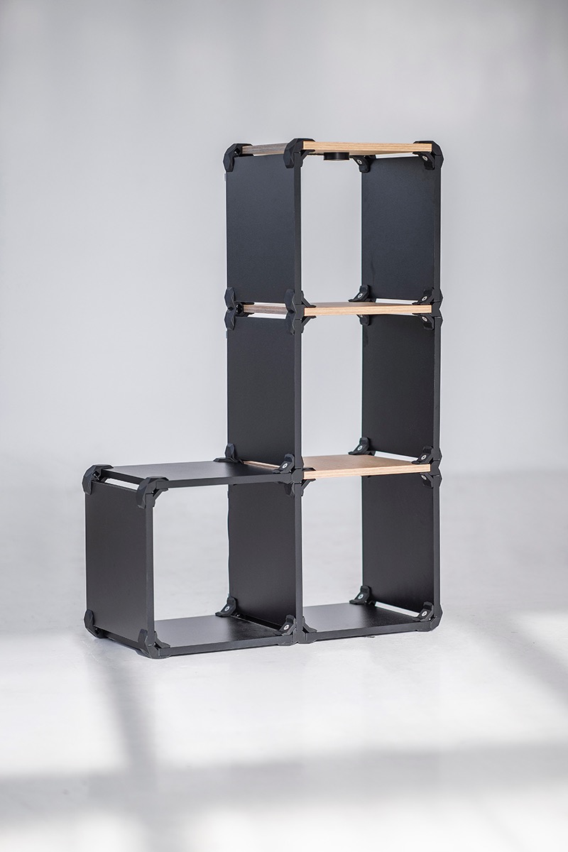 Czarny, modułowy regał z jasnymi półkami, ustawiony na białym tle. Konstrukcja złączona plastikowymi elementami, tworząca asymetryczny kształt.