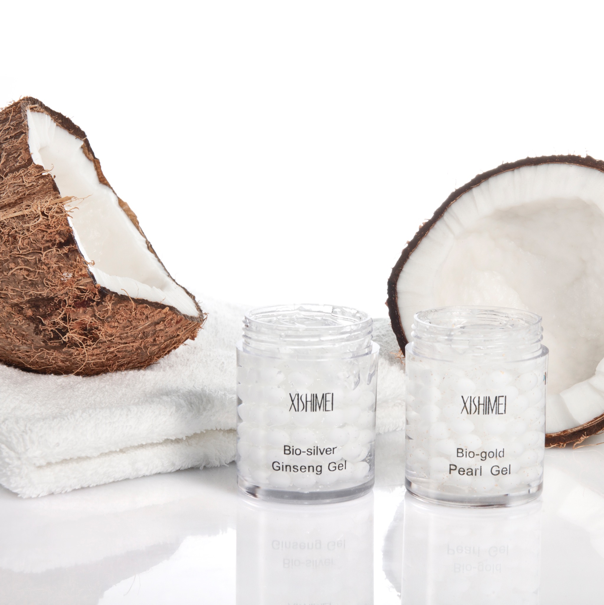 Dwa słoiczki żelu XISHIMEI (Bio-silver Ginseng Gel i Bio-gold Pearl Gel) z połówkami kokosa na białym ręczniku i błyszczącym tle. Fotografia produktowa.