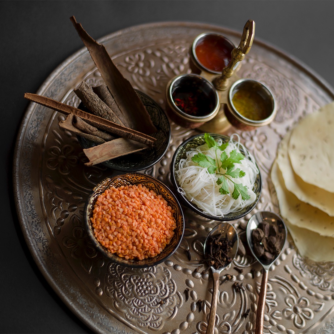 Dekoracyjna srebrna taca z miseczkami pełnymi przypraw, soczewicy, makaronu ryżowego i sosów, z dodatkiem cynamonu i przypraw na łyżeczkach.