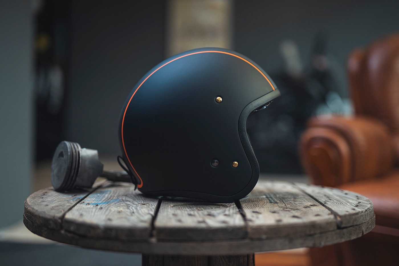 Czarny kask motocyklowy z pomarańczowym paskiem na drewnianym stoliku z elementami silnika w tle, prezentowany w studiu fotograficznym z skórzanym fotelem.