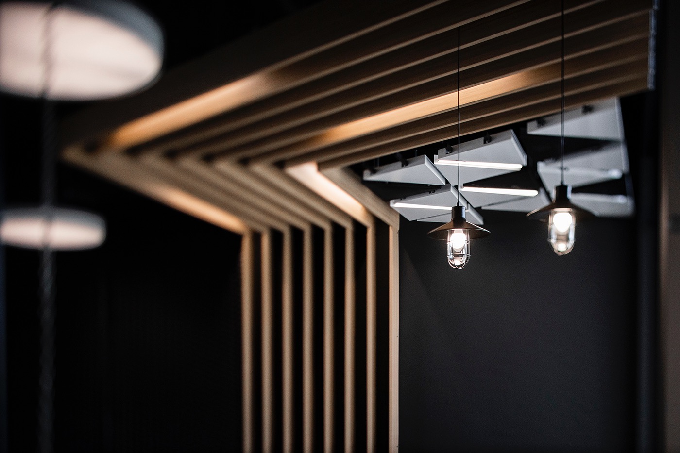 Nowoczesne oświetlenie wnętrza: geometryczny sufit z drewnianych listew, designerskie lampy wiszące z żarówkami Edisona, minimalistyczny styl i ciemne tło.