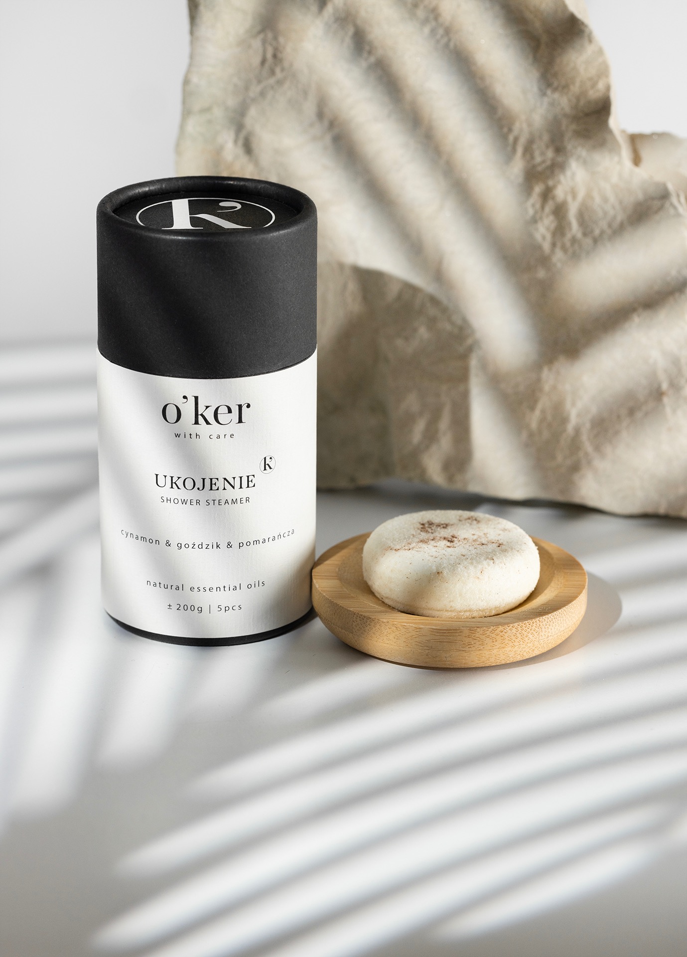 O'ker shower steamer w opakowaniu i pojedynczy steamer na drewnianej podstawce, na białym tle z cieniem liści i kamiennym tłem. Minimalistyczna fotografia produktowa.