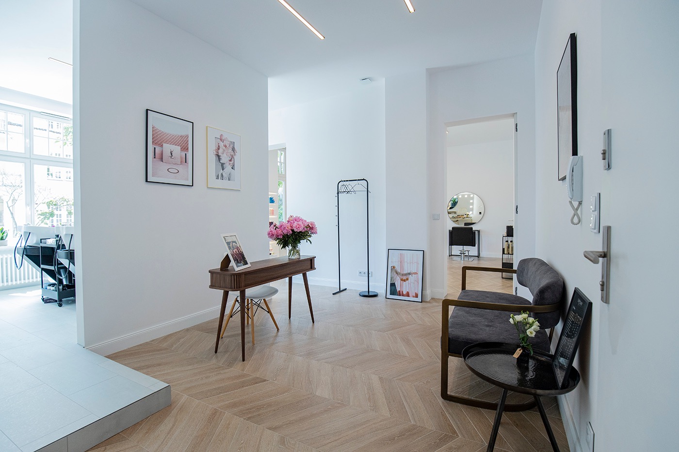 Jasne wnętrze salonu kosmetycznego z drewnianą podłogą w jodełkę, stolikiem, sofą i dekoracjami ściennymi. W tle widoczne stanowiska pracy.