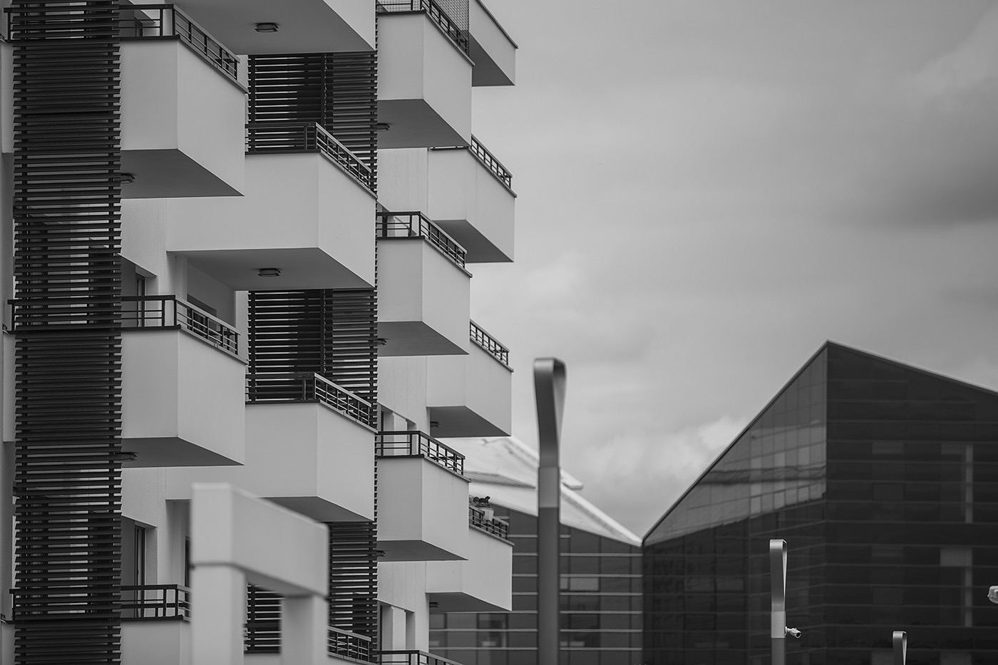 Czarno-białe zdjęcie fragmentu nowoczesnego budynku mieszkalnego z balkonami, z widocznym budynkiem biurowym w tle i elementami oświetlenia ulicznego.