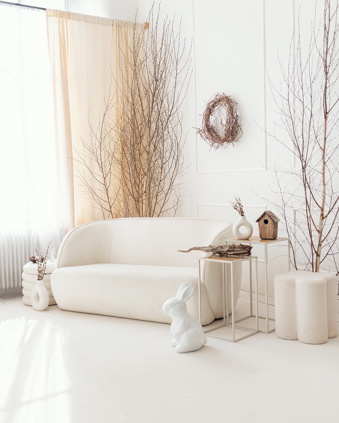 Jasne wnętrze z kremową sofą, dekoracjami z gałęzi, białym królikiem i minimalistycznymi stolikami. Wieniec na ścianie dodaje naturalnego akcentu.
