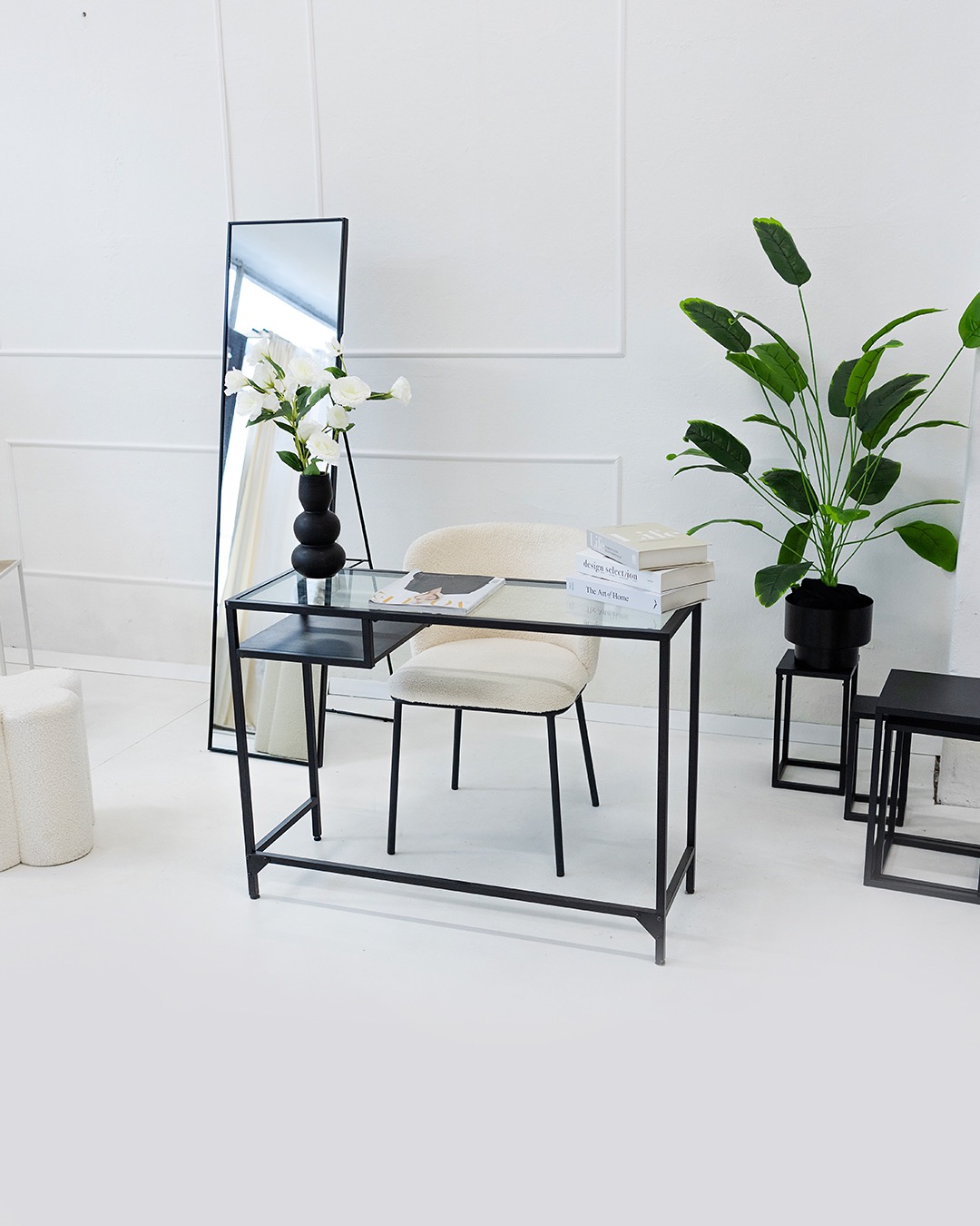 Minimalistyczne wnętrze: szklane biurko z czarną ramą, kremowe krzesło, lustro w czarnej ramie, kwiaty w wazonie i roślina doniczkowa na tle białej ściany.