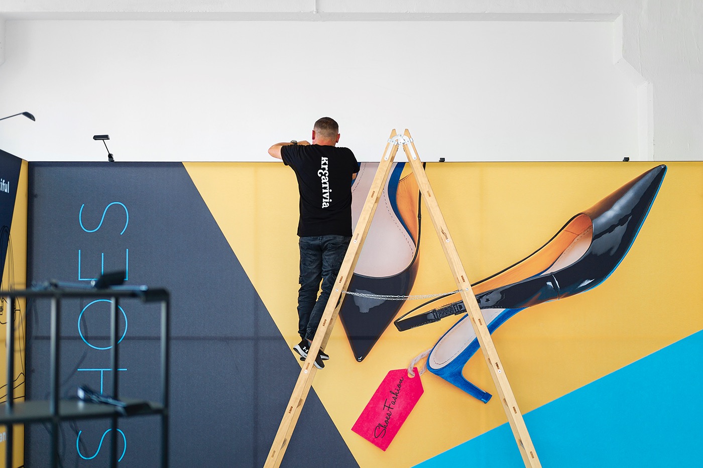 Mężczyzna na drabinie montuje duży baner reklamowy z grafiką butów na tle żółto-niebieskim. Baner w trakcie instalacji, widoczne narzędzia i drabina.