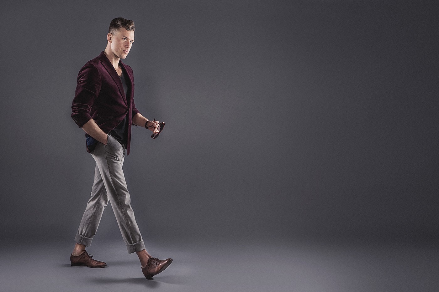 Mężczyzna w bordowej marynarce i szarych spodniach, idący na szarym tle, trzymający okulary w dłoni.