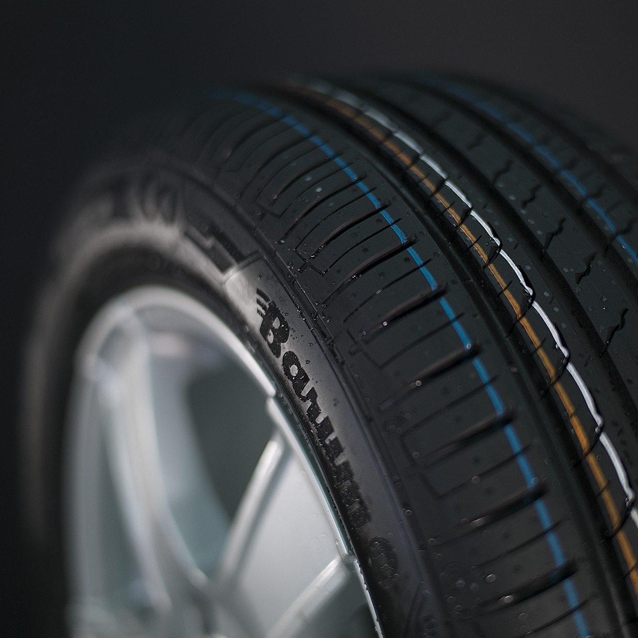 Precyzyjne ujęcie opony samochodowej z aluminiową felgą, podkreślone detale bieżnika z kolorowymi liniami i logo producenta w lekko wilgotnym wykończeniu na ciemnym tle.