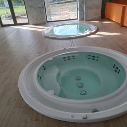 ROBERT KIEŁBASA - Dwie okrągłe wanny z hydromasażem, jedna z widocznymi dyszami i bąbelkami, umieszczone na drewnopodobnej podłodze w jasnym wnętrzu z dużymi oknami i elementami dekoracyjnymi.