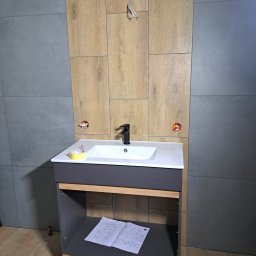 ROBERT KIEŁBASA - Półka pod umywalkę z czarną baterią, zamontowana na tle ściany z szarych i drewnopodobnych płytek, z widocznymi puszkami instalacyjnymi i instrukcją montażu na półce