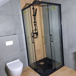 ROBERT KIEŁBASA - Nowoczesna kabina prysznicowa z czarnymi profilami i deszczownicą, drewnopodobne panele ścienne za kabiną, szare płytki na pozostałych ścianach, wisząca toaleta.