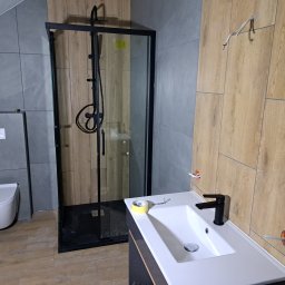 ROBERT KIEŁBASA - Łazienka w trakcie wykańczania: czarna kabina prysznicowa typu walk-in, biała umywalka z czarną armaturą, płytki imitujące drewno na ścianie, widoczne przewody elektryczne i taśma malarska.