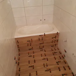 Układanie płytek imitujących drewno na podłodze i częściowo na ścianie w łazience, z użyciem klipsów poziomujących, widoczna wanna zabezpieczona folią.