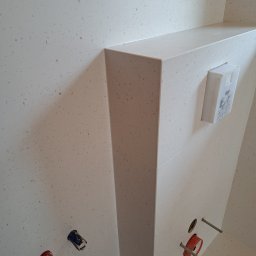 ROBERT KIEŁBASA - Ściana łazienki wyłożona jasnymi płytkami z drobnymi brązowymi plamkami, widoczne elementy instalacji sanitarnej i stelaż podtynkowy Grohe.