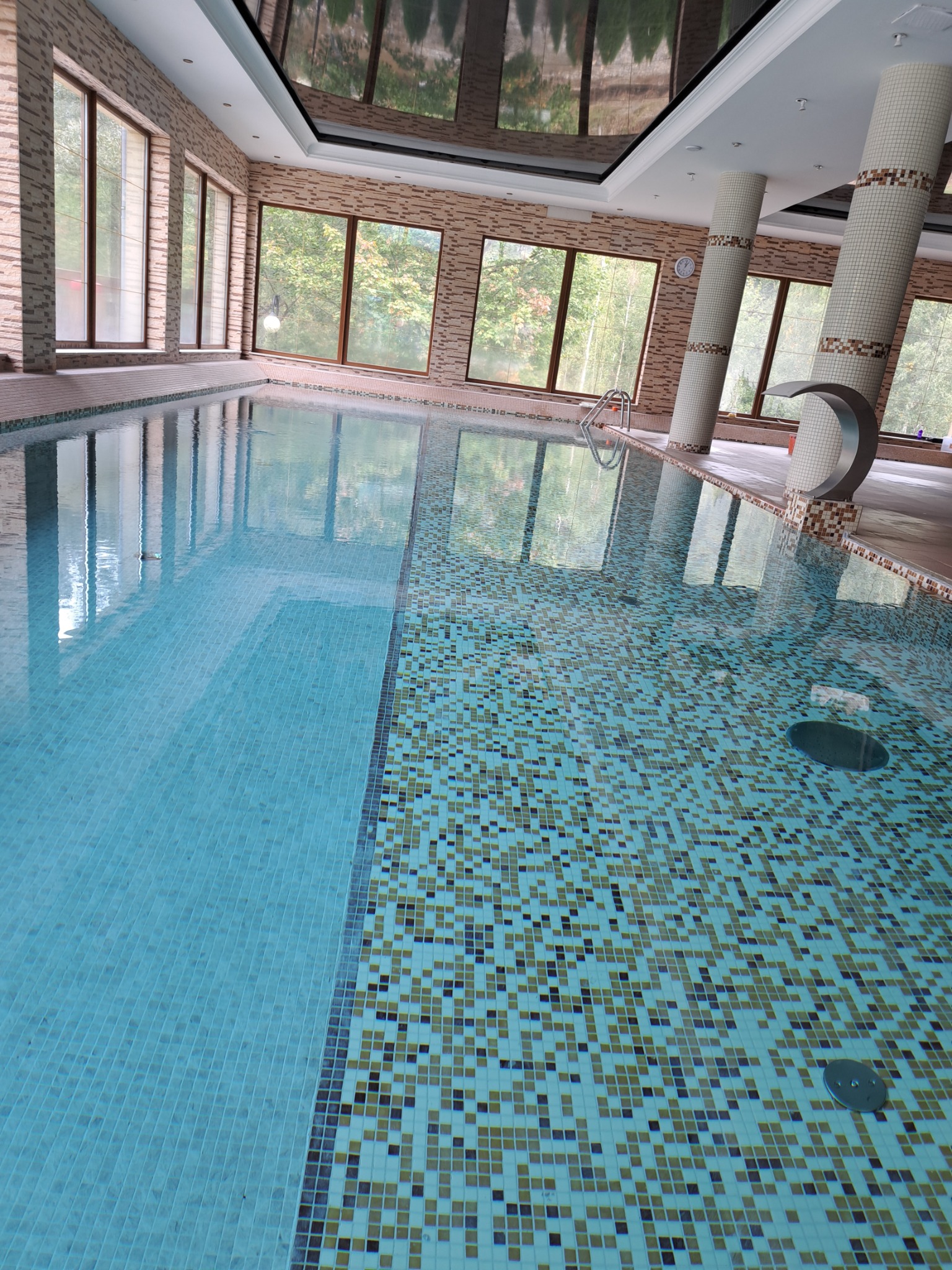 Kryty basen z mozaiką w Nysie, widok na kolumny z dekoracyjną mozaiką i okna z widokiem na drzewa, woda oświetlona naturalnym światłem.