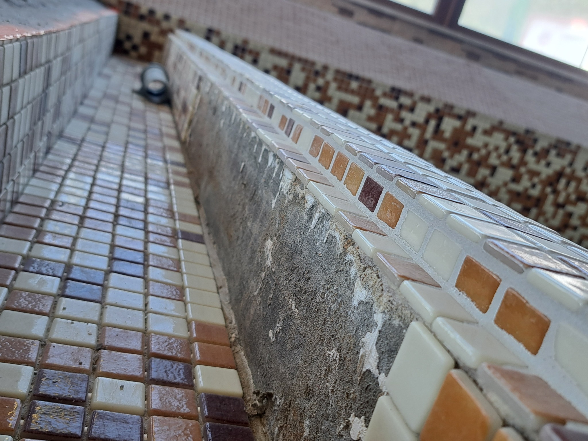 Nisko ustawiony kadr na fragment wykończonej mozaiką ściany łazienki, z widocznym fragmentem okna w tle i niedokładnościami w położeniu płytek mozaiki przy krawędzi ściany.