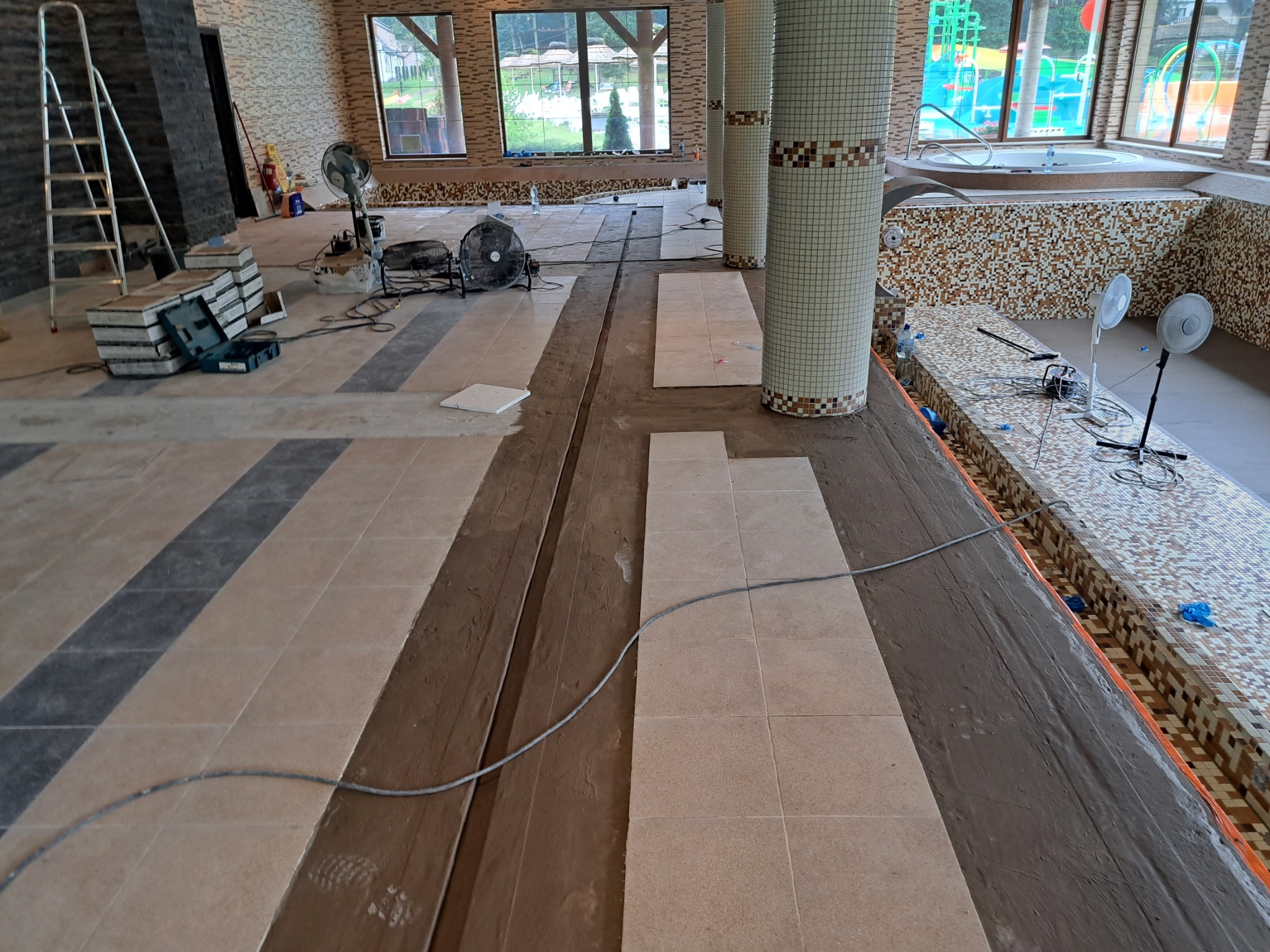 Wnętrze w trakcie układania jasnych płytek podłogowych wzdłuż basenu z mozaikową obudową, widoczne kolumny wykończone mozaiką, drabina, wentylatory i narzędzia glazurnicze.