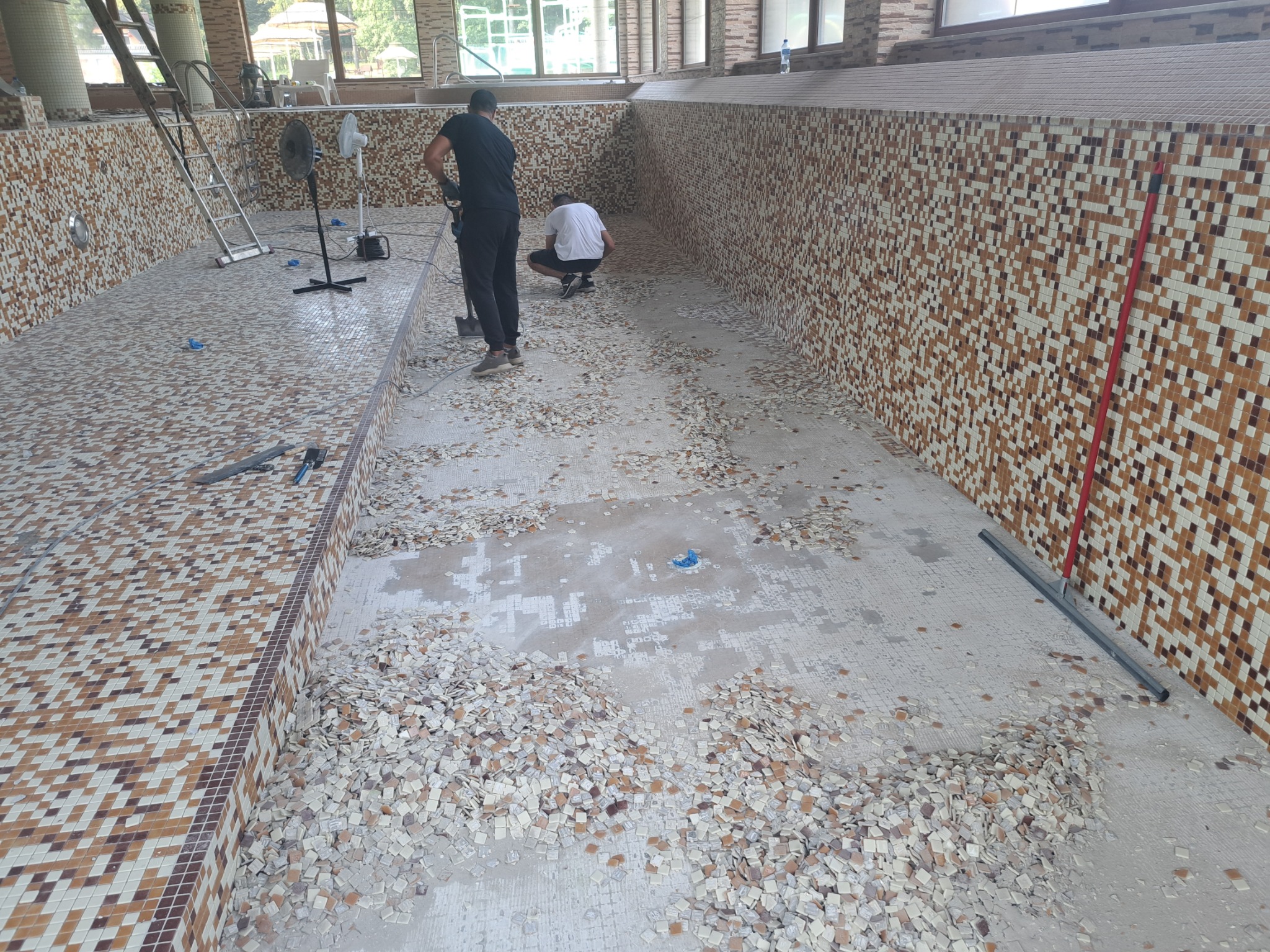 Demontaż mozaiki w pustym basenie wewnętrznym; widoczne odspojone płytki, pracownicy z narzędziami, drabina i wentylatory.