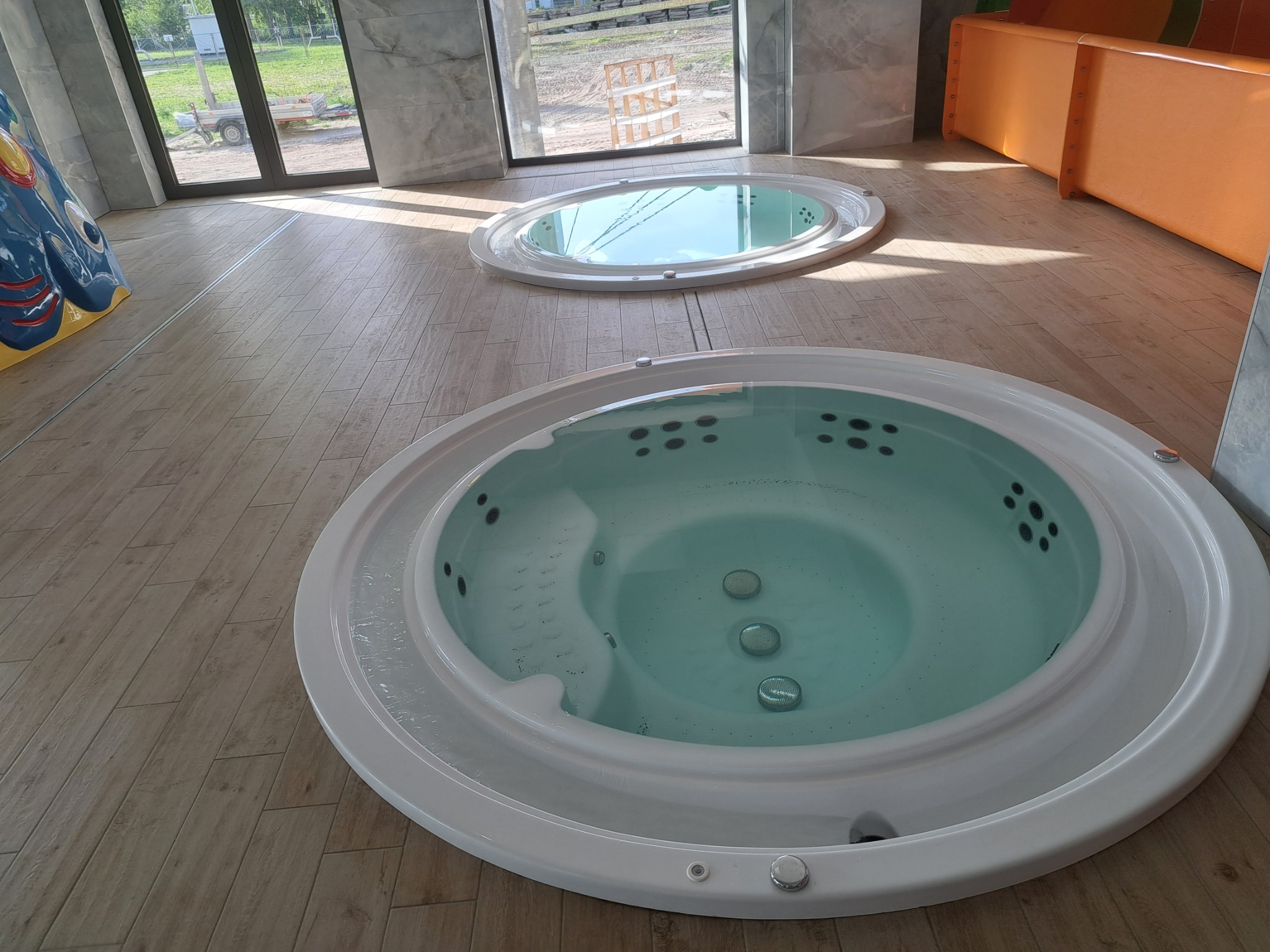 Dwie okrągłe wanny z hydromasażem, jedna z widocznymi dyszami i bąbelkami, umieszczone na drewnopodobnej podłodze w jasnym wnętrzu z dużymi oknami i elementami dekoracyjnymi.