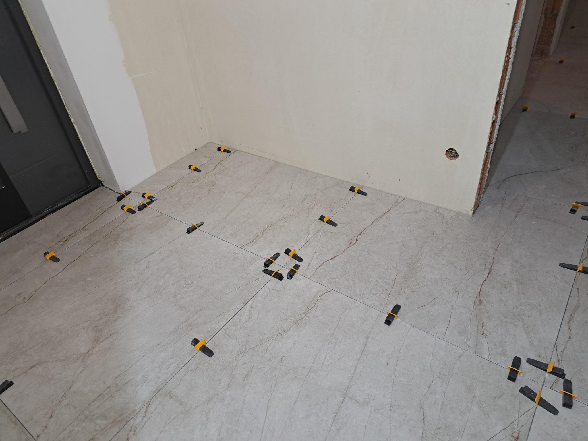 Układanie płytek podłogowych z użyciem czarnych klipsów poziomujących z żółtymi klinami w narożnikach, widoczny fragment ściany z niedokończonym malowaniem.