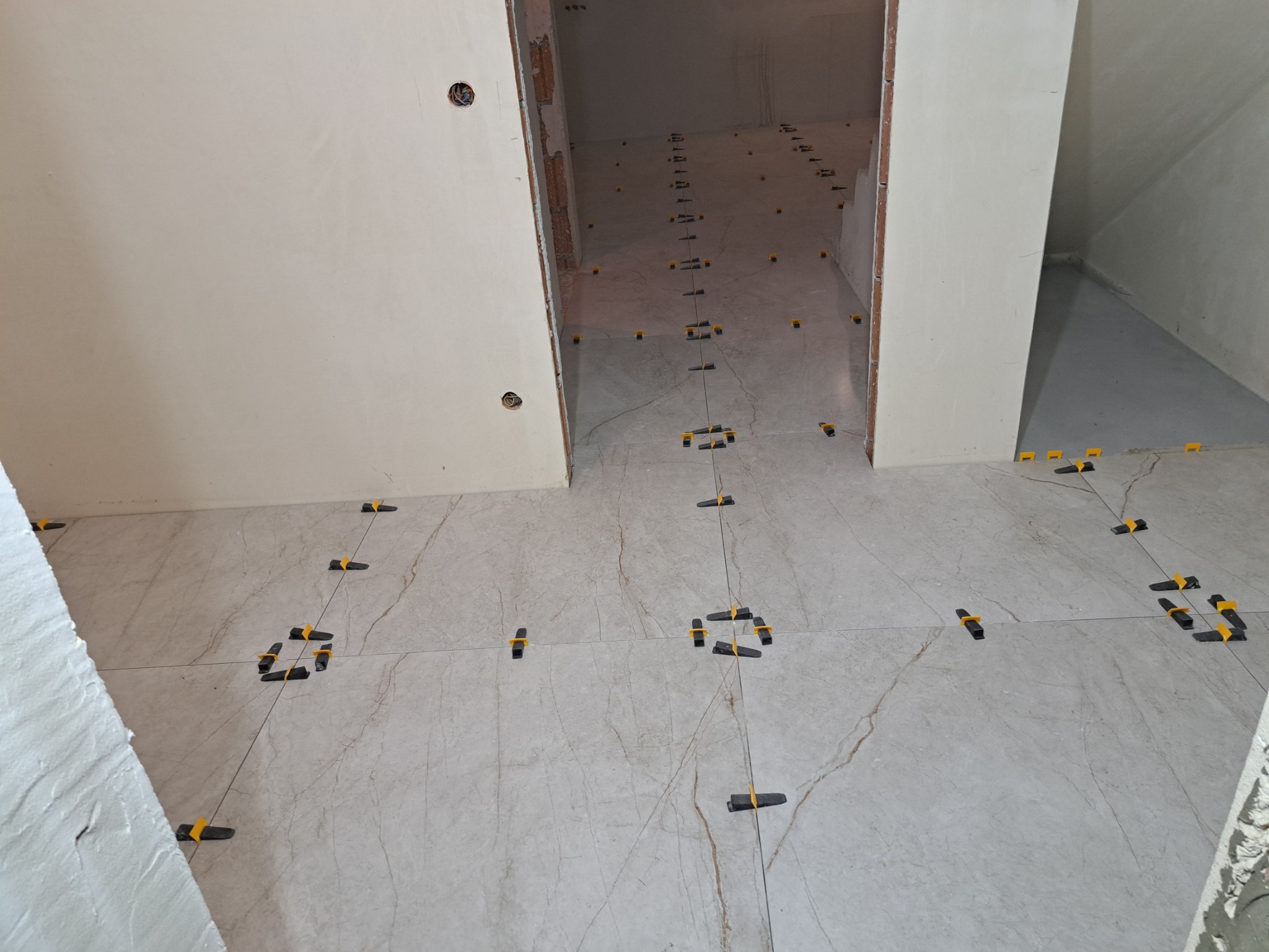 Układanie marmurowych płytek podłogowych z użyciem systemu poziomowania, widoczne czarne klipsy z żółtymi klinami i sznurki, surowe ściany w tle.