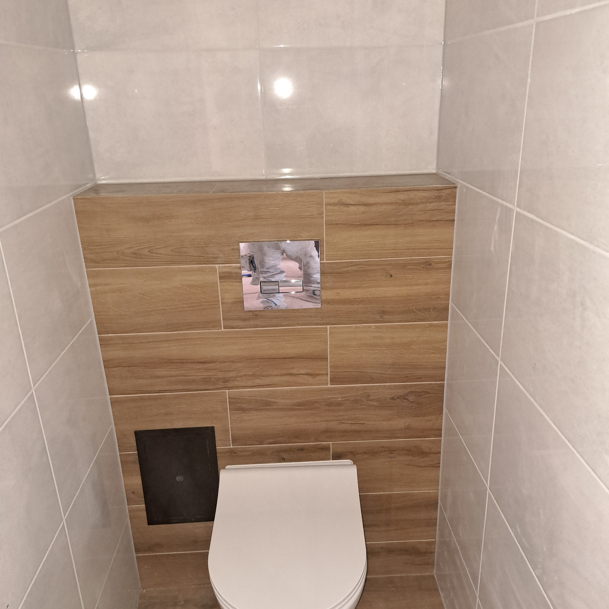 Wykończona toaleta w łazience z płytkami imitującymi drewno i jasnymi płytkami ceramicznymi, widoczna klapa rewizyjna i chromowany przycisk spłuczki.