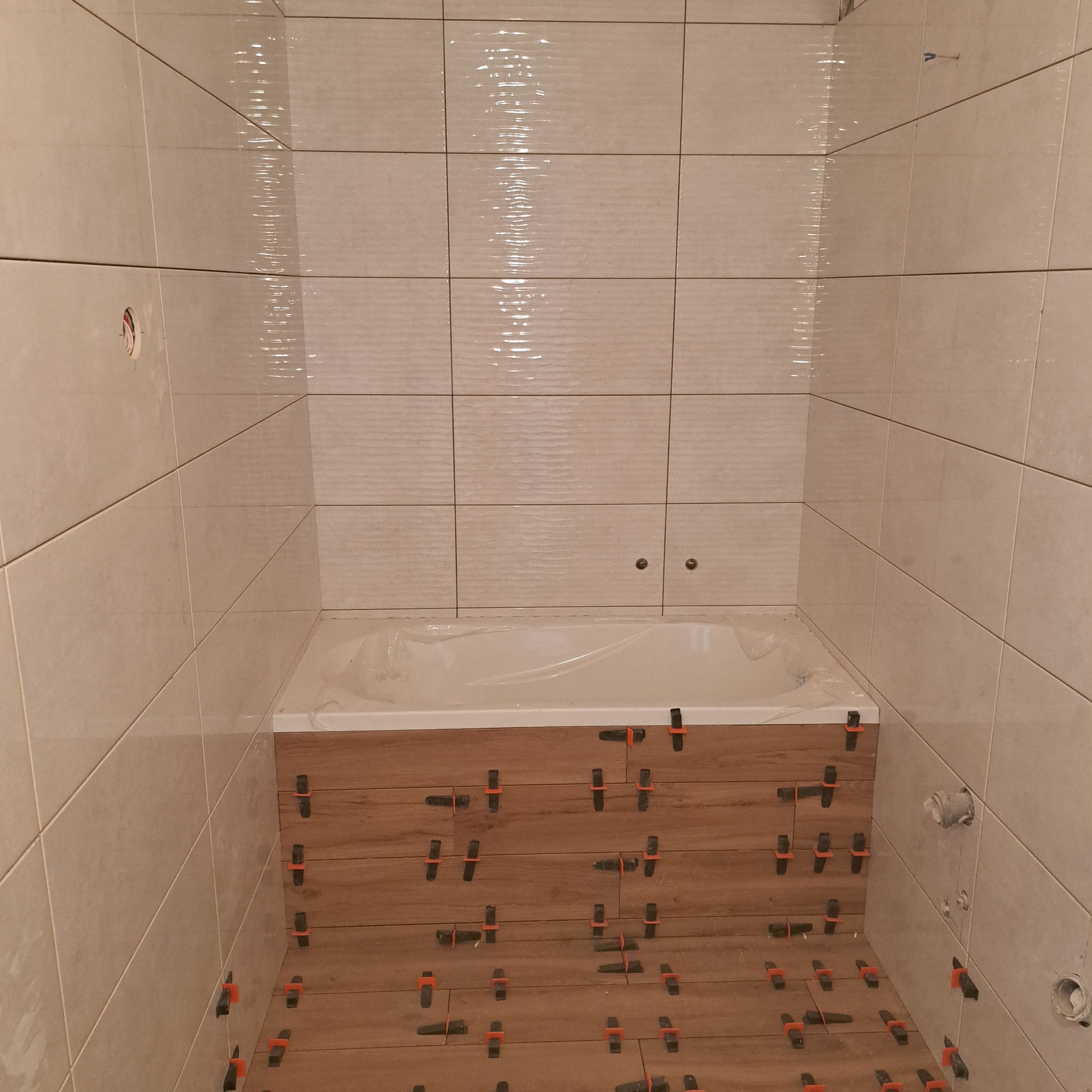 Układanie płytek imitujących drewno na obudowie wanny, jasne płytki ścienne z fakturą, plastikowe krzyżyki dystansowe w spoinach.