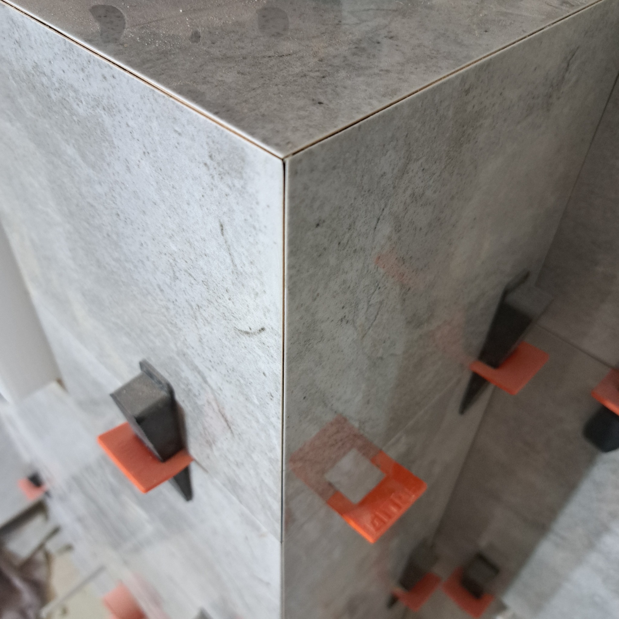 Fragment narożnika obłożonego szarymi płytkami imitującymi kamień, z widocznymi pomarańczowymi i czarnymi klipsami poziomującymi płytki.