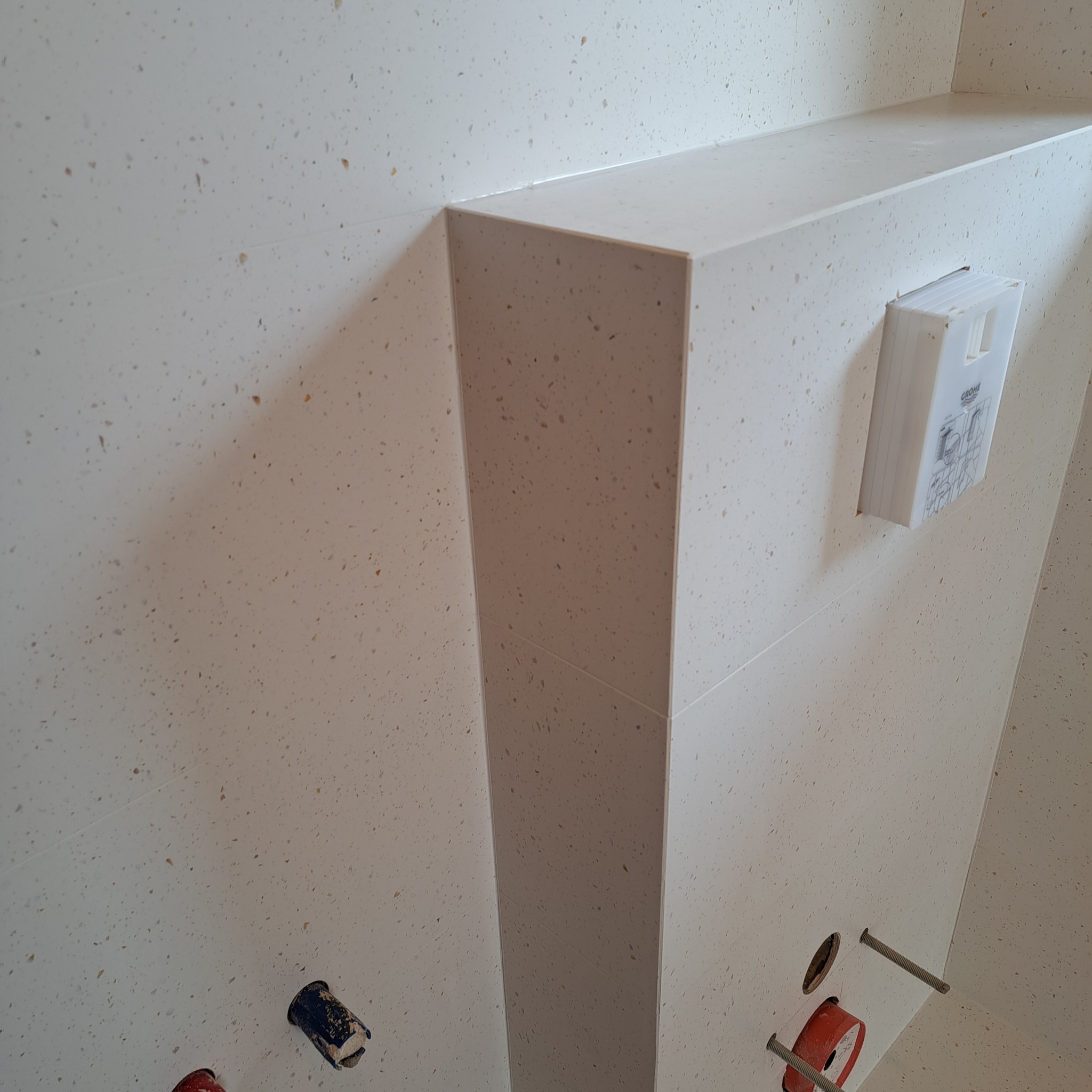 Ściana łazienki wyłożona jasnymi płytkami z drobnymi brązowymi plamkami, widoczne elementy instalacji sanitarnej i stelaż podtynkowy Grohe.