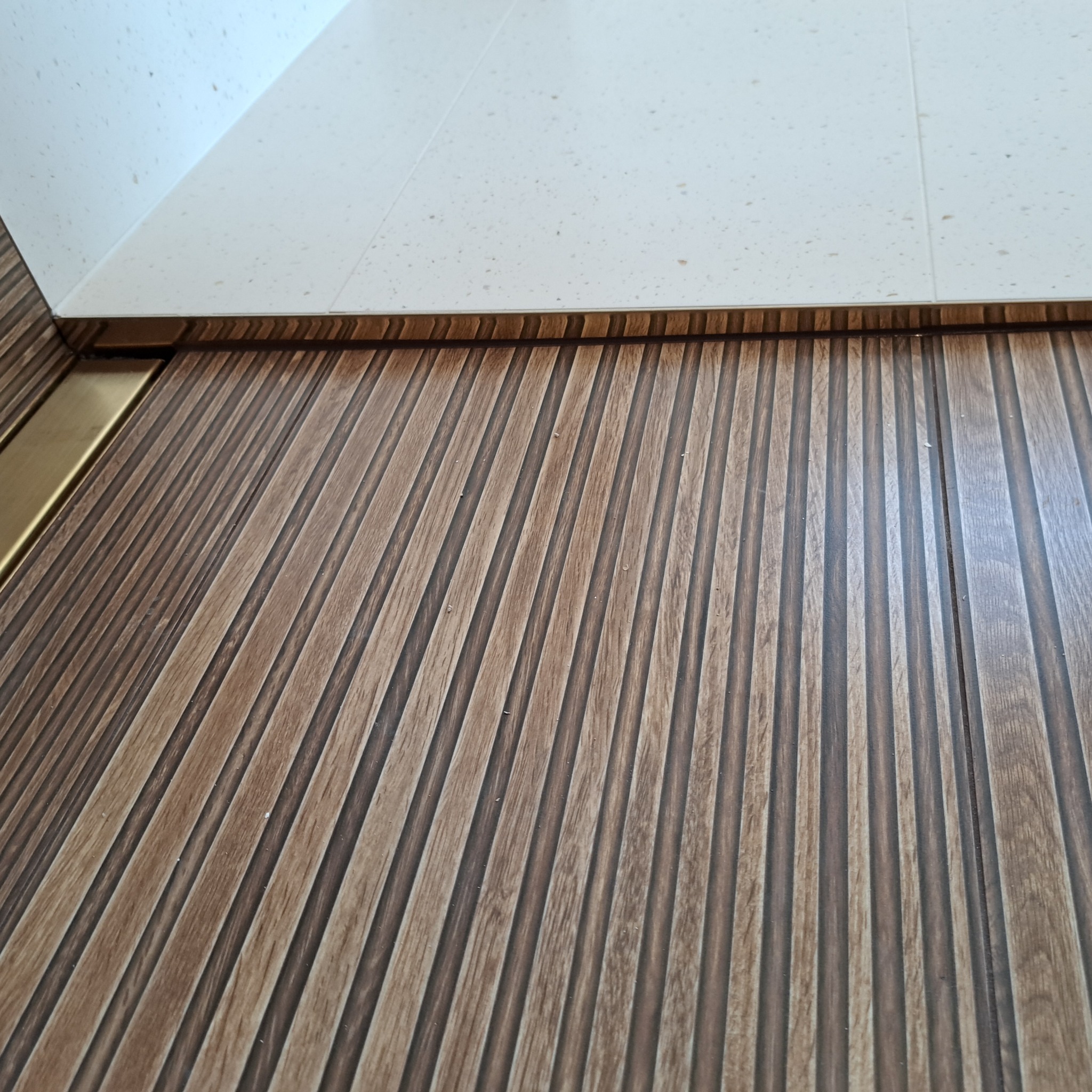 Detaliczne ujęcie podłogi w łazience z drewnopodobnych paneli z wyraźnymi, pionowymi żłobieniami, przylegającej do jasnych, kwadratowych płytek na ścianie, oddzielonych złotą listwą wykończeniową.