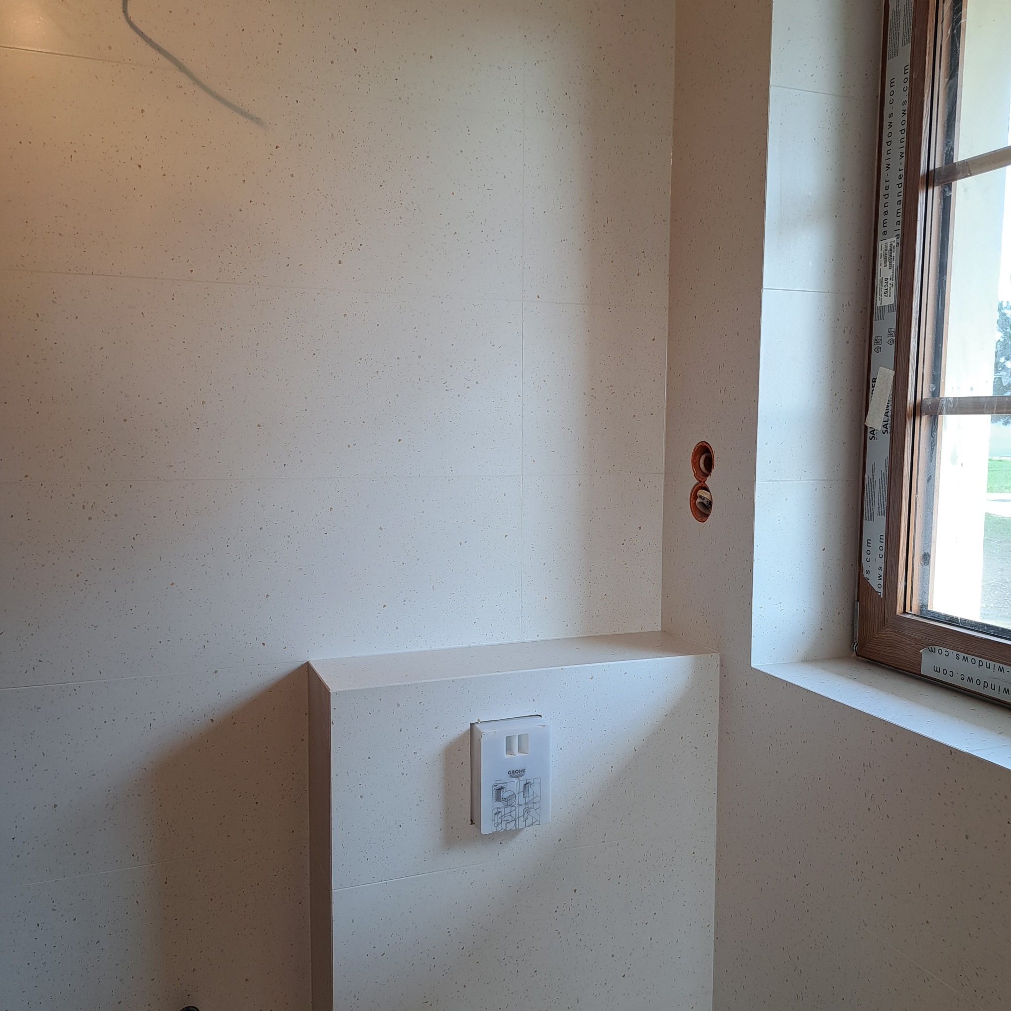 Ściana łazienki wyłożona jasnymi płytkami z drobnym wzorem, widoczne otwory na instalację elektryczną i przycisk spłuczki WC marki Grohe, fragment okna z drewnianą ramą.