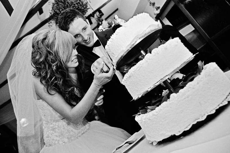 Czarno-biała fotografia pary młodej krojącej trzypiętrowy tort weselny, panna młoda w sukni z tiulowym welonem, pan młody w smokingu z muchą, uśmiechnięci podczas uroczystości.