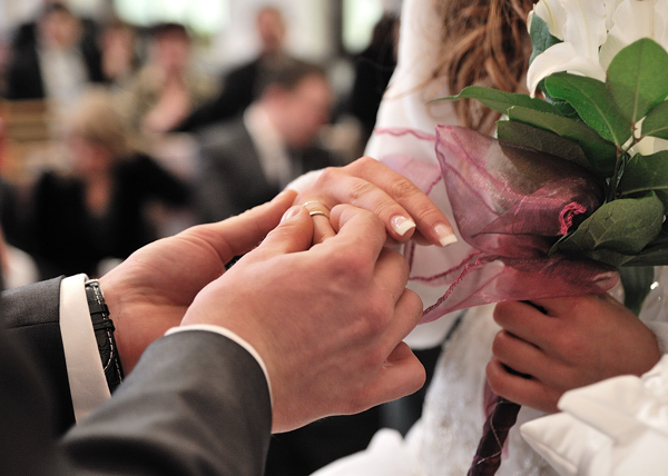 Moment zakładania złotej obrączki na palec panny młodej podczas ceremonii ślubnej, bukiet ślubny w tle.