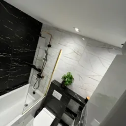 Nowoczesna łazienka z czarnymi i białymi marmurkowymi płytkami, złotymi akcentami na armaturze i kabinie prysznicowej, minimalistyczna toaleta i umywalka, oraz roślina doniczkowa.