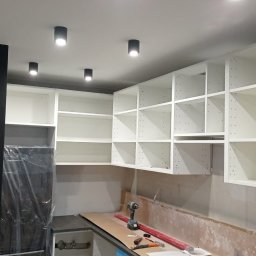 Wojtek Turowski - Montaż białych szafek kuchennych z widoczną wiertarką i poziomicą na blacie, czarny front lodówki zabezpieczony folią.